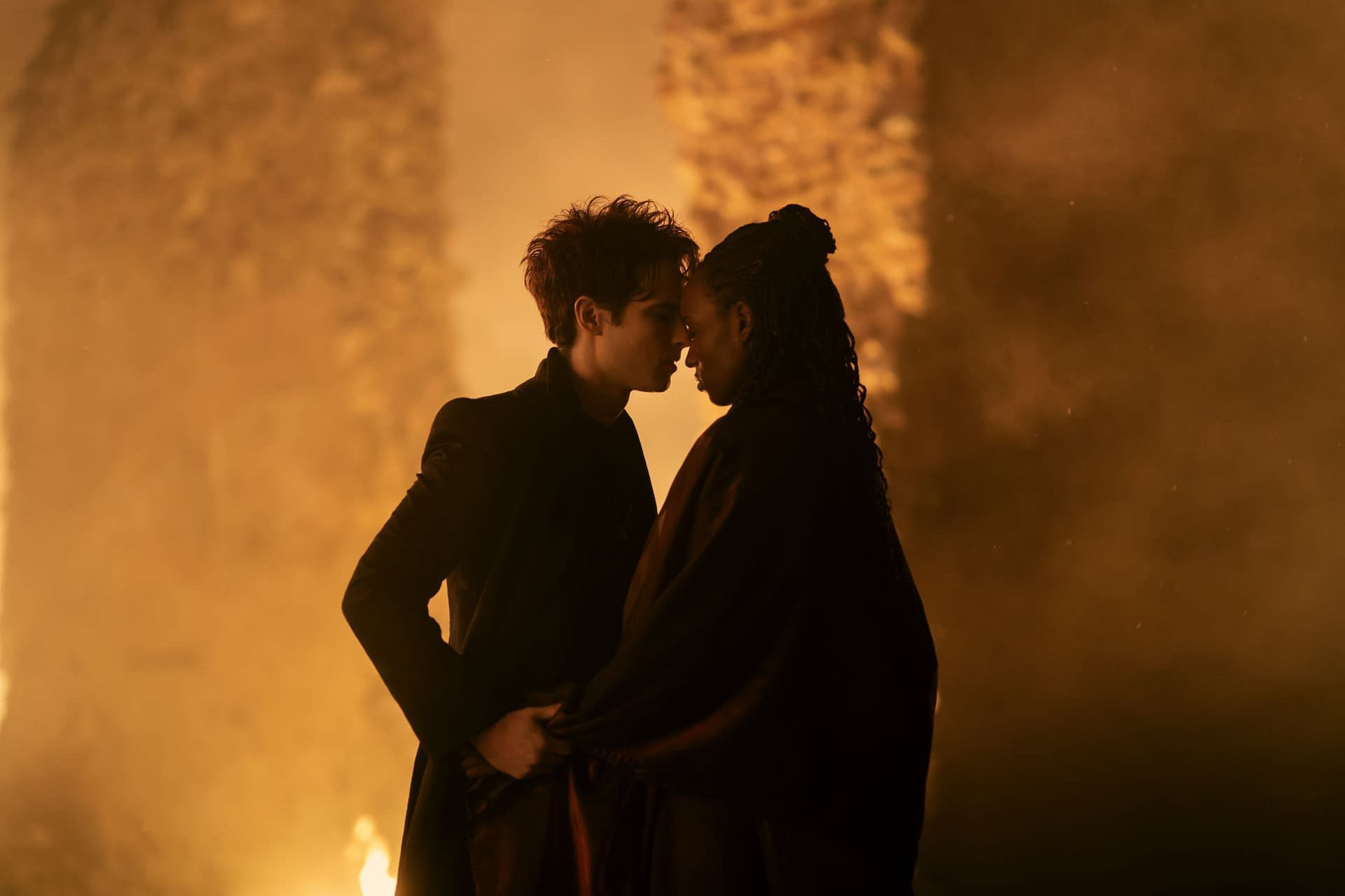 Tom Sturridge as Dream (Morpheus) and Deborah Oyelade as Queen Nada share an intimate moment, standing forehead to forehead against a fiery, ancient backdrop in The Sandman (Netflix series).